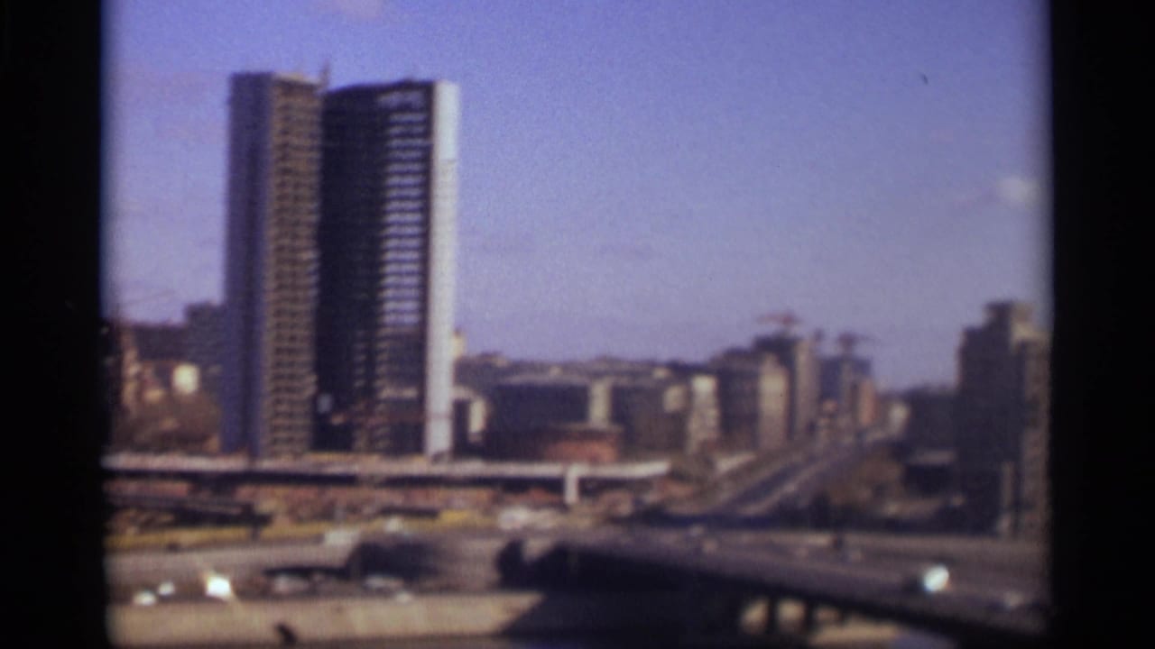 Cars cross a bridge over water with a tall building and city skyline beyond.