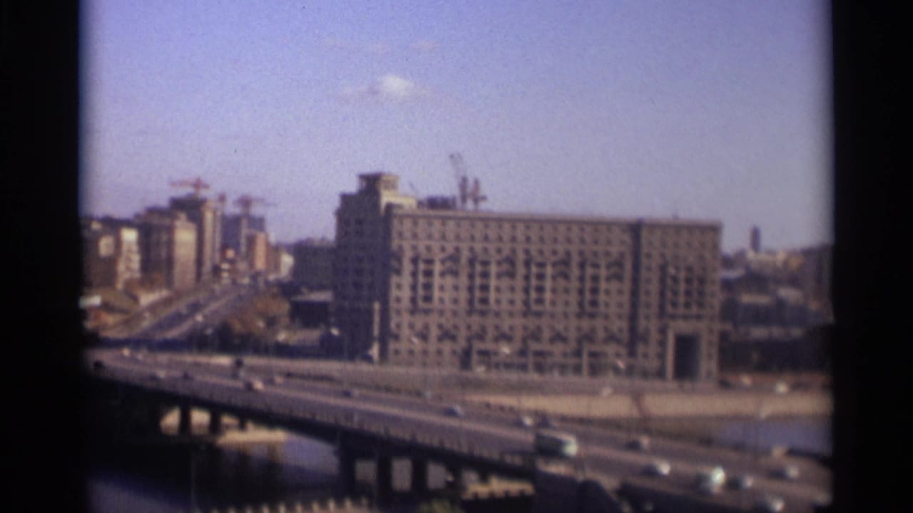 Bridge traffic crossing a river with a large gridded building and cranes beyond.