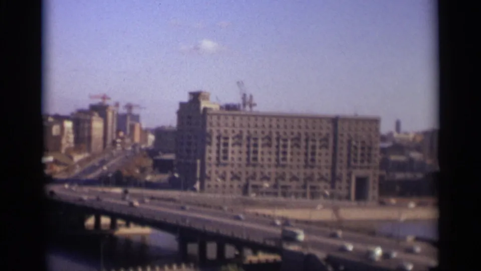 1966 Moscow home-movie frame showing cars on a broad bridge, river water, and a large riverside building with rooftop cranes.