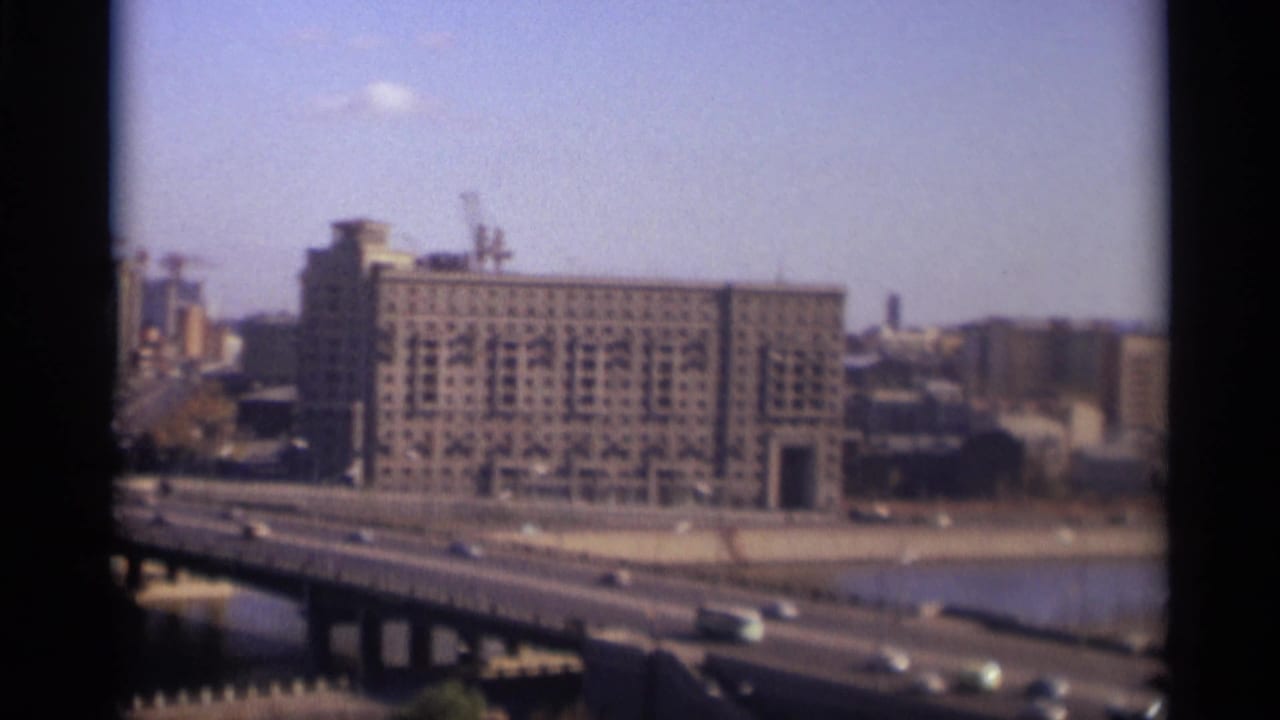 River bridge and traffic with a large building, cranes, and city blocks in the background.