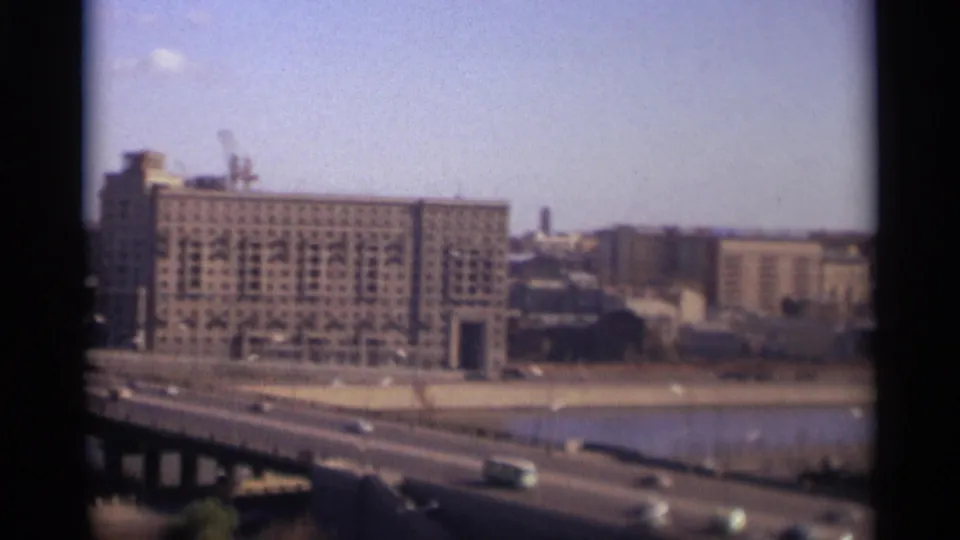 1966 Moscow view of a large gridded riverside building, construction cranes, nearby blocks, and cars crossing a bridge in front.