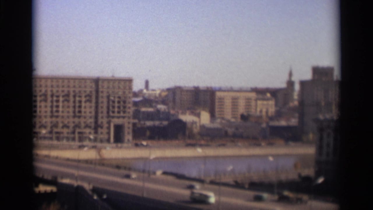 1966 Moscow home-movie frame showing a riverfront roadway, moving cars, and large Soviet-era buildings in soft focus.