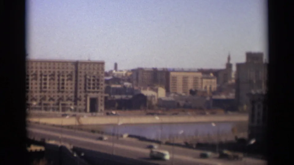 1966 Moscow home-movie frame showing a riverfront roadway, moving cars, and large Soviet-era buildings in soft focus.