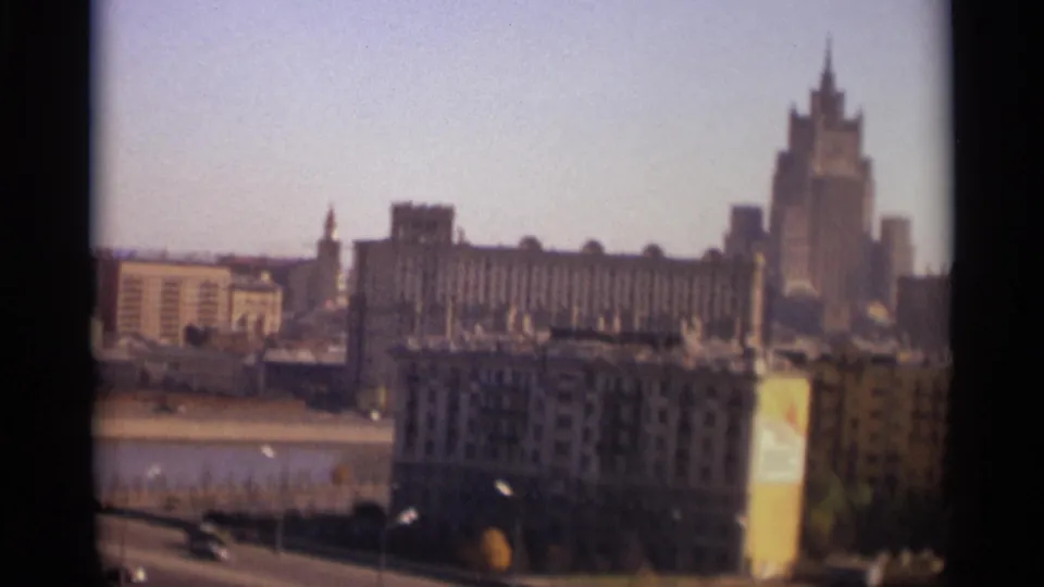 Moscow riverfront skyline with a broad road below, small moving cars, apartment blocks, and a prominent tower in the distance.