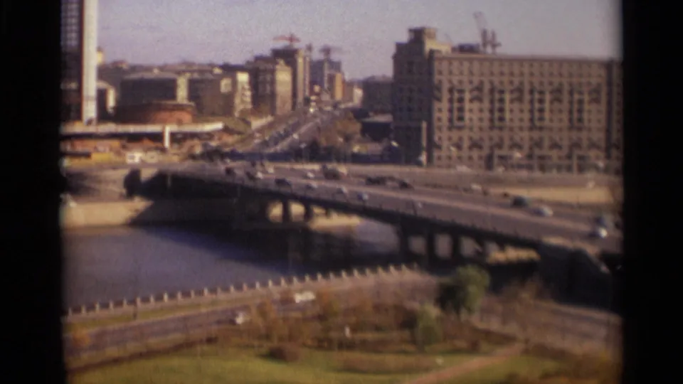 1966 Moscow river bridge scene with moving cars, a large riverside block, landscaped embankment, and cranes in the distance.