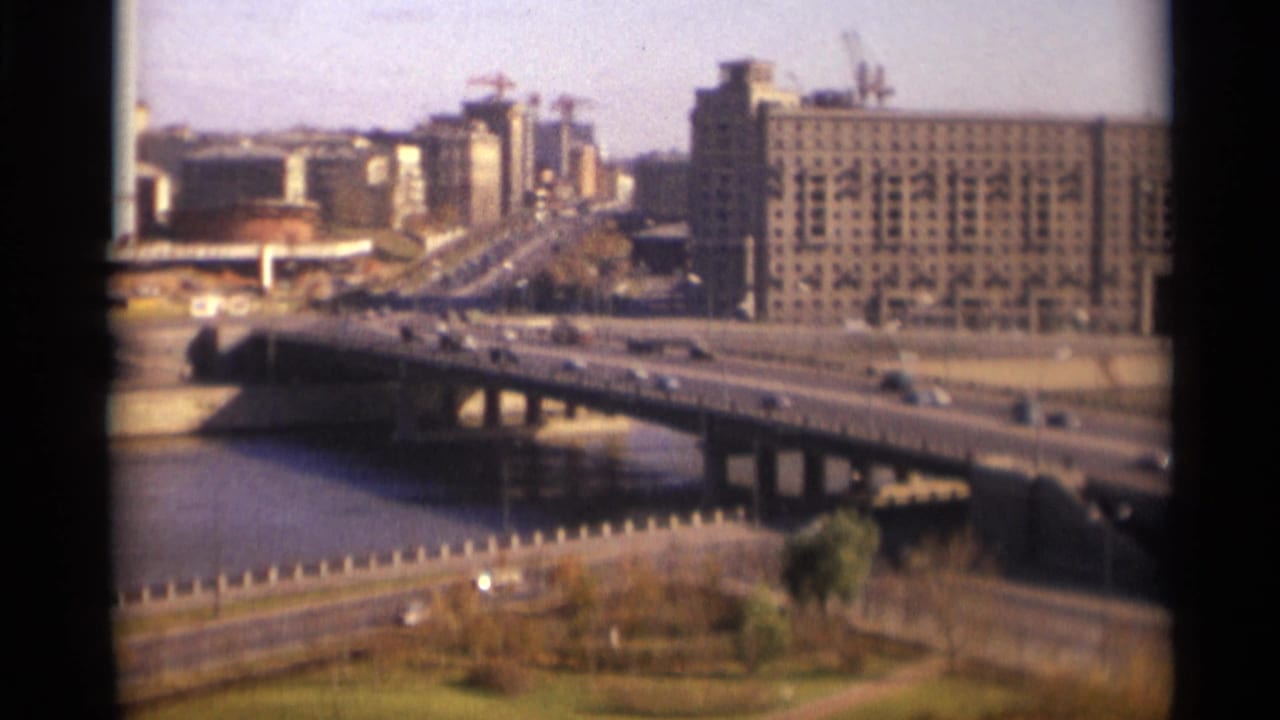 Elevated view of cars crossing a broad bridge over a river with large buildings beyond.