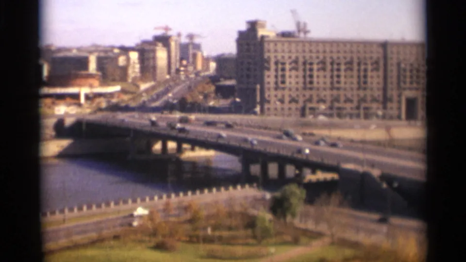 Soft 8mm frame from 1966 Moscow showing cars on a broad bridge above the river, with large urban buildings and cranes behind it.