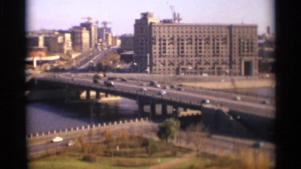 1966 Moscow frame showing a river bridge with passing cars, foreground parkland, embankment railings, and large buildings beyond.