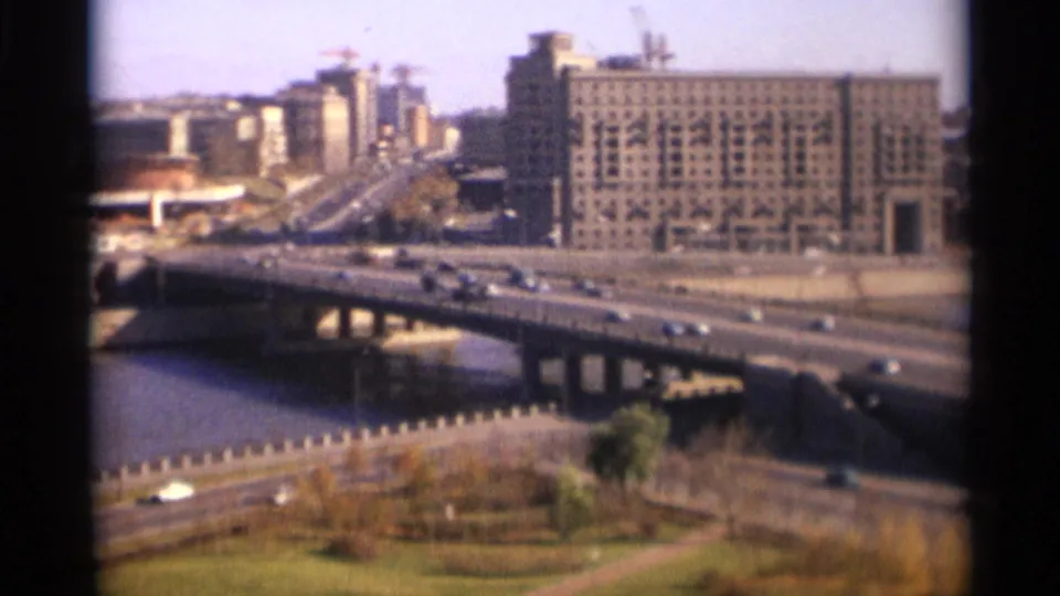 Faded 8mm view of 1966 Moscow traffic crossing a river bridge, framed by dark film edges and distant Soviet-era urban blocks.