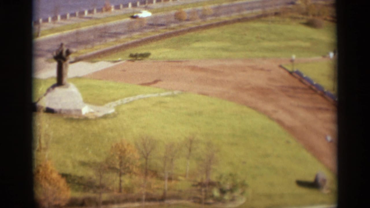1966 Moscow home-movie frame showing an elevated park view with a monument on a stone base, lawns, road, and distant traffic.