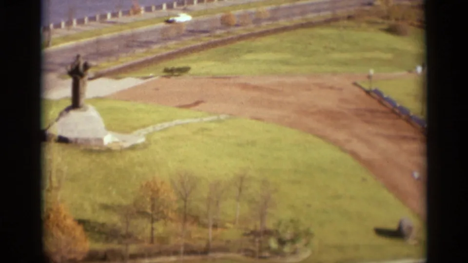 1966 Moscow home-movie frame showing an elevated park view with a monument on a stone base, lawns, road, and distant traffic.