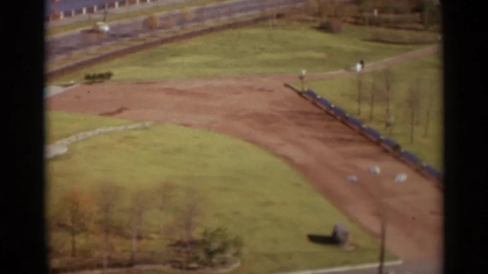 Elevated 1966 Moscow park frame showing lawns, a reddish path, roadside edge, sparse trees, and distant people in the grounds.