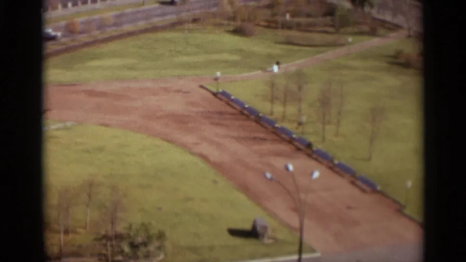 1966 Moscow home-movie frame showing an elevated view over a dirt park path lined with blue benches and sparse trees.