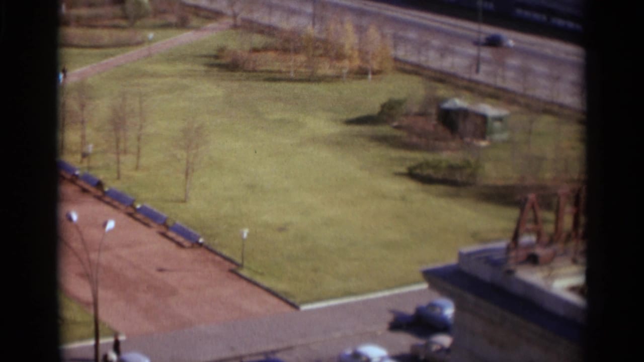 Elevated view of grassy urban grounds, paths, parked cars, and construction activity at frame edge.