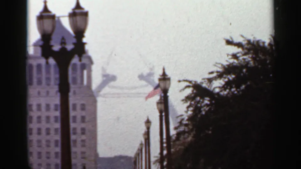 1966 Saint Louis home-movie view with ornate streetlamps, a tall building, American flag, and the Gateway Arch faintly visible in haze.