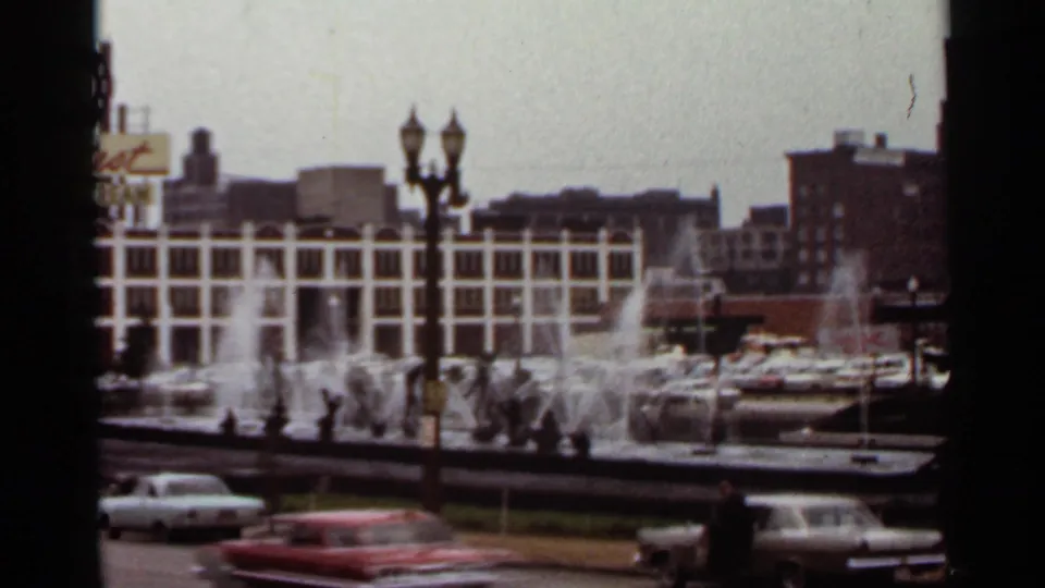 1966 Saint Louis plaza scene with active fountain jets, classic cars, streetlamps, and mid-century buildings around the square.