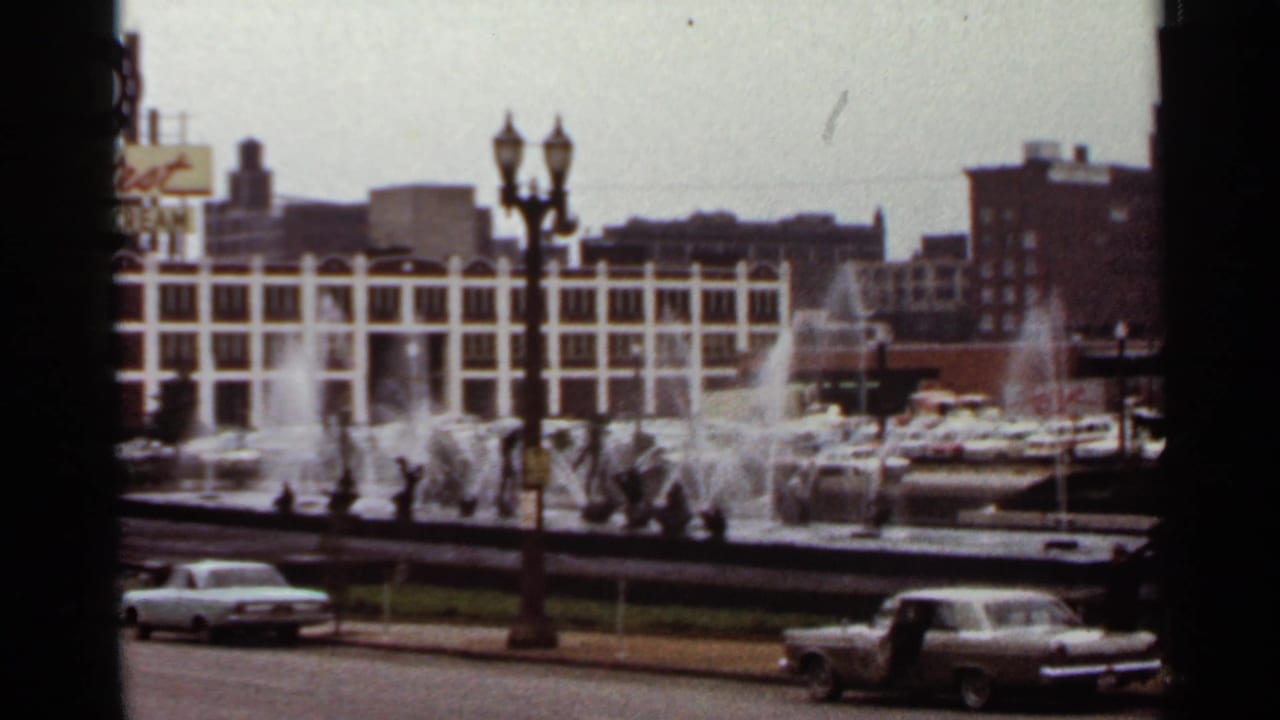 Fountains spray beside street lamps as vintage cars pass a city street.