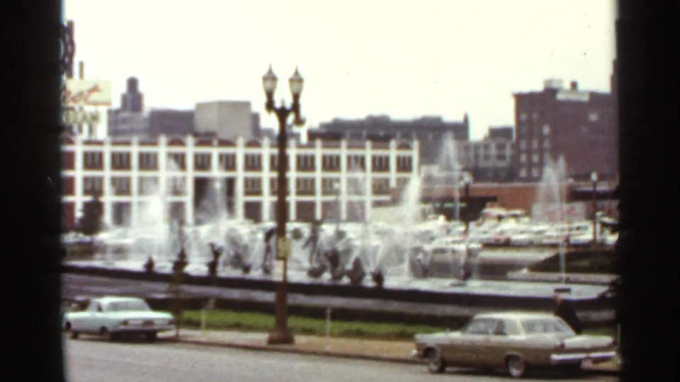Saint Louis fountain plaza with water jets, ornamental lamps, older cars, and a white building facade, filmed on 8mm in 1966.