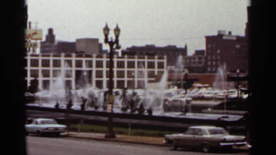 Wide 8mm view of a Saint Louis fountain area, street lamps, period cars, and downtown buildings in faded color.