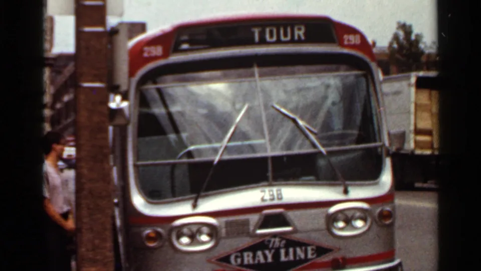 Close view of a red and silver Gray Line tour bus in Saint Louis, with partial pedestrians and a truck nearby in 1966 home-movie footage.