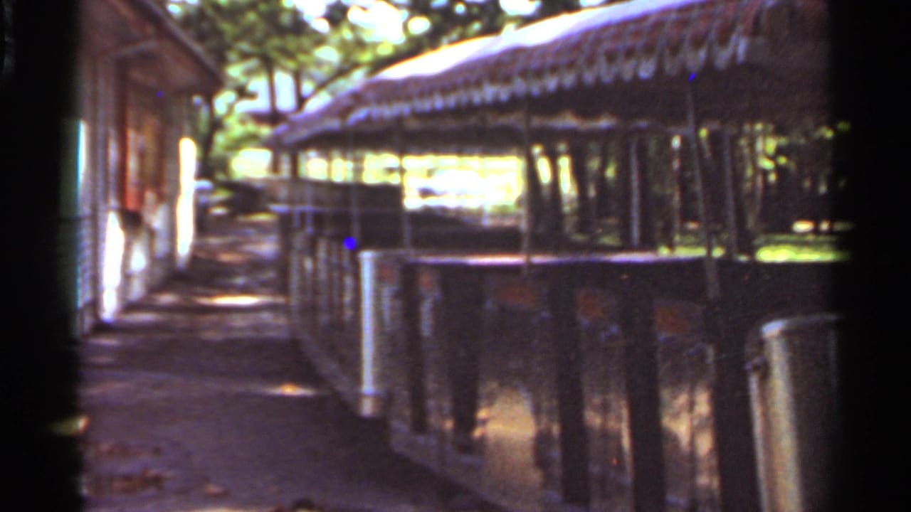 1966 Saint Louis home-movie frame showing open-sided amusement park train cars near a shaded path and small building.