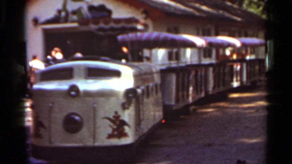 1966 Saint Louis home-movie frame showing a small amusement park locomotive with passenger cars near a mural-fronted building.