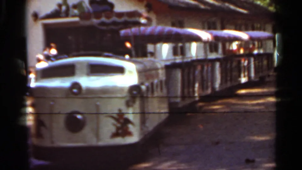 1966 Saint Louis frame of a cream amusement train passing with purple-striped canopy cars behind it.