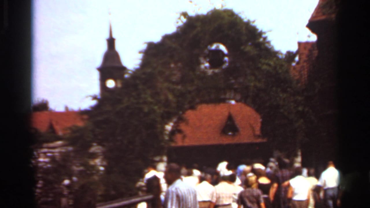 1966 Saint Louis home-movie frame showing visitors approaching a green archway with a clock tower and red-tiled roof in the background.