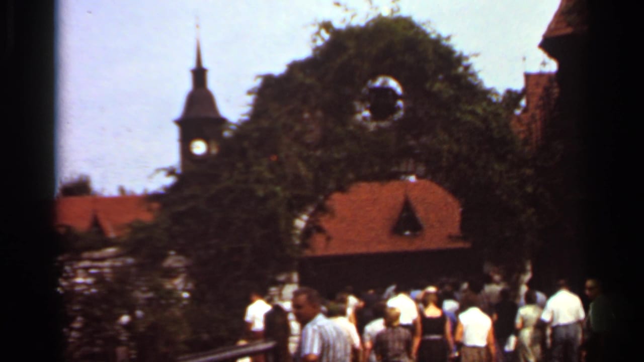 1966 Saint Louis frame of adults walking through a green archway near a stone wall, with a red roof and clock tower beyond.