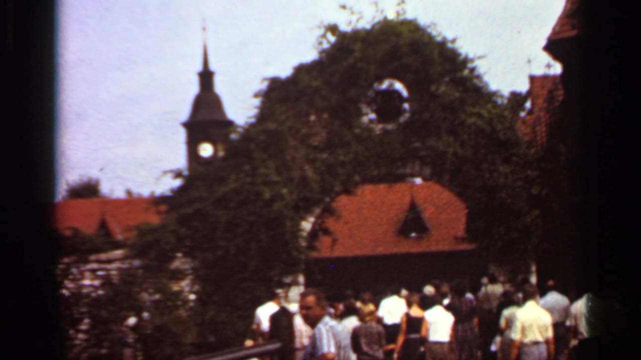 1966 Saint Louis home-movie view of visitors clustered near a vine-covered arch, red-tiled roof, and distant clock tower.