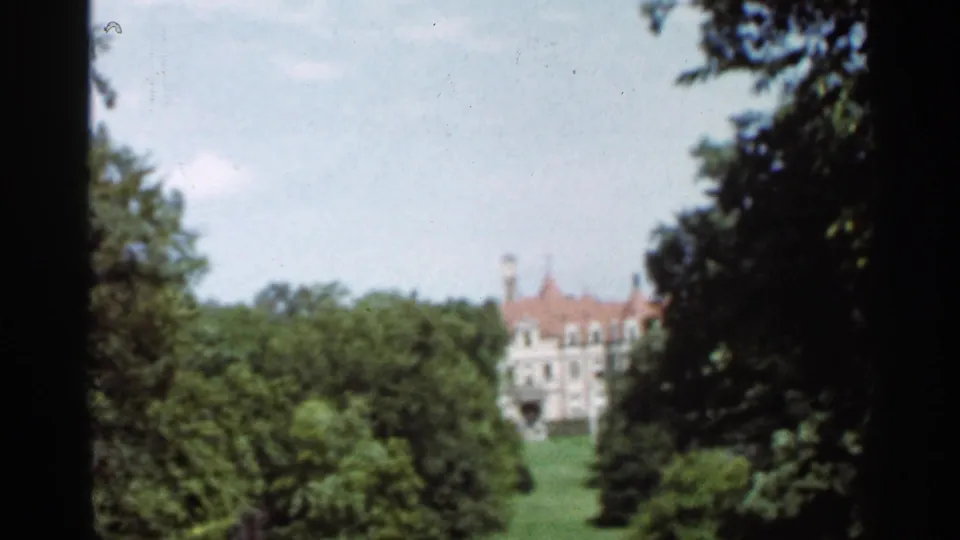 Vintage 8mm frame from Saint Louis showing a landscaped green slope, trees, and a distant light-colored building.