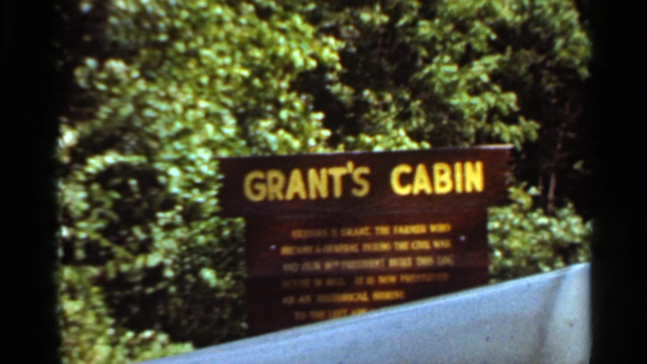 Wooden Grant's Cabin sign partly blocked by a pale foreground barrier, with dense green trees behind it.
