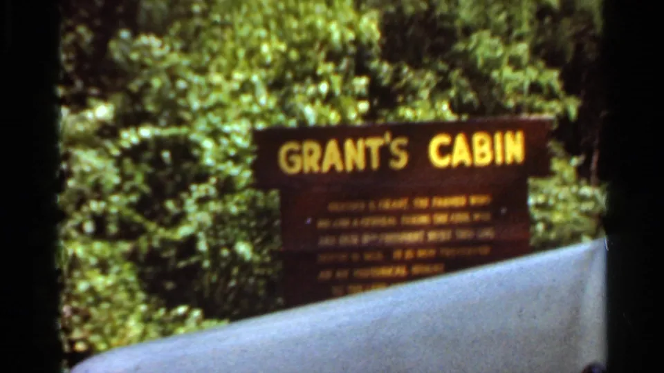 Visitor-shot 8mm view of the Grant's Cabin sign in Saint Louis, with yellow lettering, surrounding trees, and a pale foreground barrier across the lower frame.