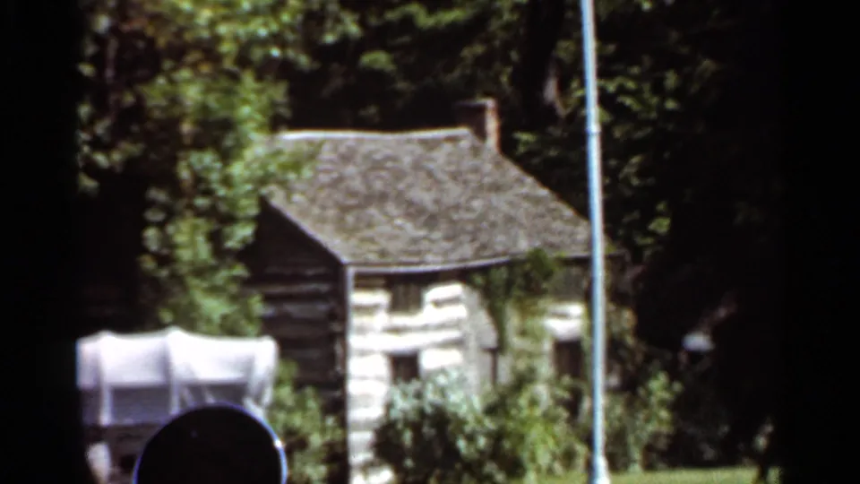 1966 Saint Louis home-movie frame showing Grant's Cabin with a shingled roof and chimney, plus a white covered wagon, flagpole, grass, and surrounding trees.