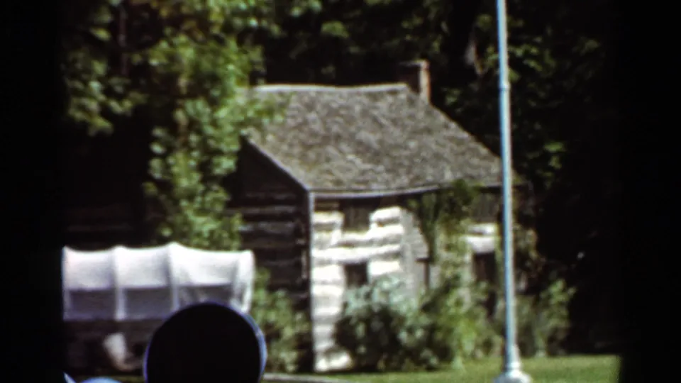 Soft 8mm view of Grant's Cabin in Saint Louis, showing the log structure, covered wagon, flagpole, lawn, trees, and dark frame edges.