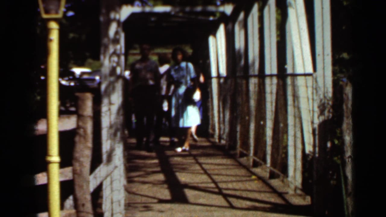 In 1966 Saint Louis home-movie footage, two adults approach along a wooden walkway beneath a white metal structure with a yellow post at left.