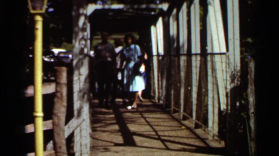 In 1966 Saint Louis home-movie footage, two adults approach along a wooden walkway beneath a white metal structure with a yellow post at left.