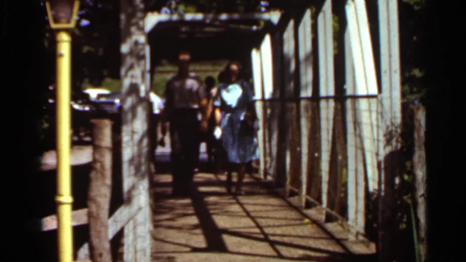 The pair move closer on the Saint Louis walkway, with metal framing, railing shadows, a yellow post and parked cars visible beyond the bridge.