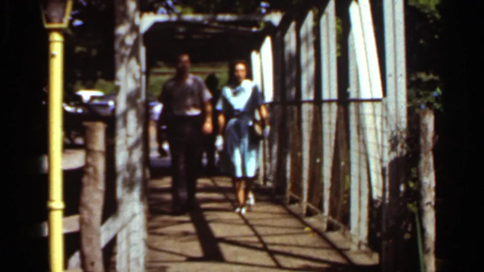 Two adults in mid-1960s clothing walk side by side toward the camera beneath the white structure, with bright sun and dark rail shadows across the deck.