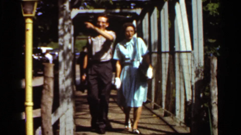 The older man points or gestures while walking with the woman on the bridge in 1966 Saint Louis home-movie footage.