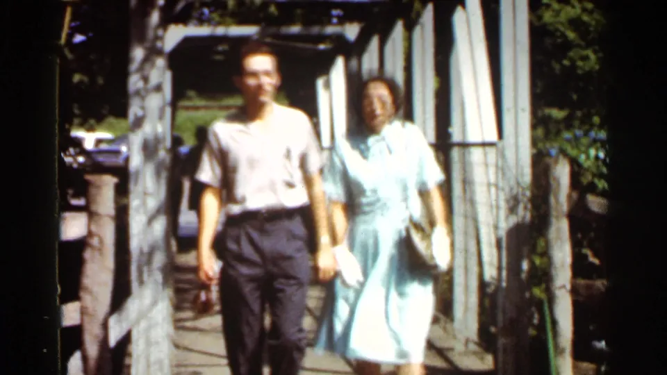 The couple nears the camera, holding hands on the wooden bridge, with strong sunlight and 8mm color softness.