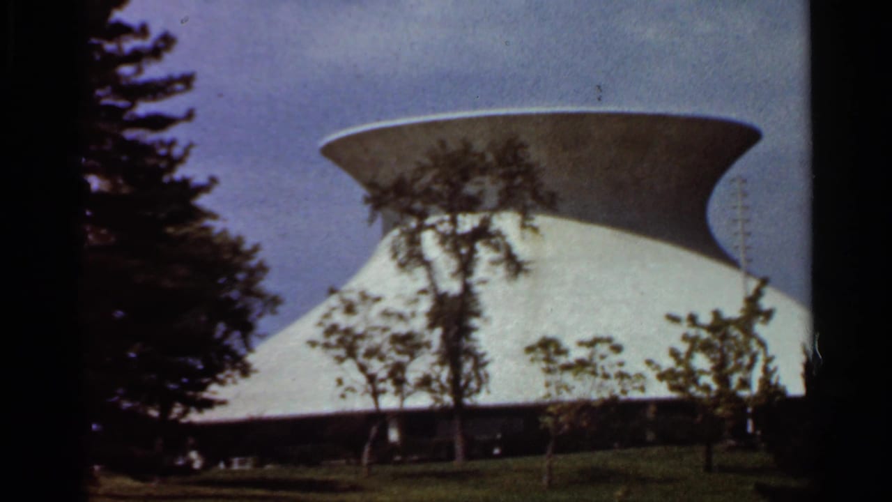 White modernist building with a flared circular roof partly framed by trees.