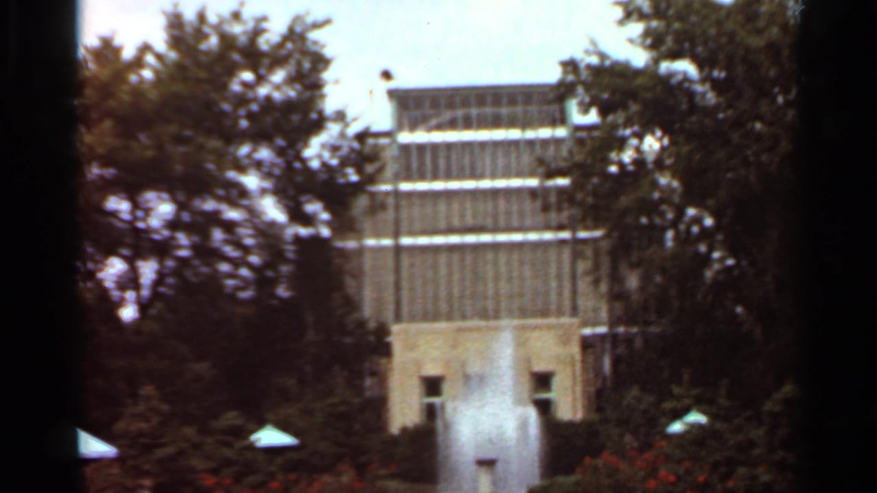 1966 Saint Louis home-movie frame showing a fountain jet before a modernist building, trees, low plantings, and dark film edges.