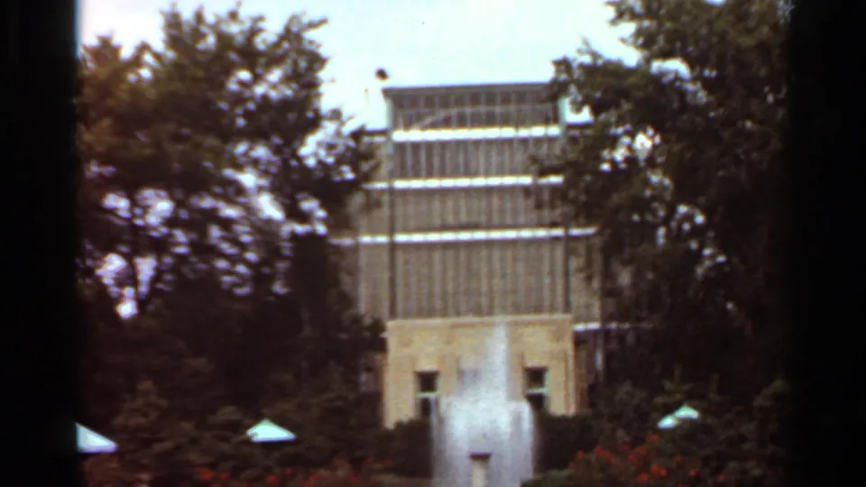 1966 Saint Louis home-movie frame showing a fountain jet before a modernist building, trees, low plantings, and dark film edges.