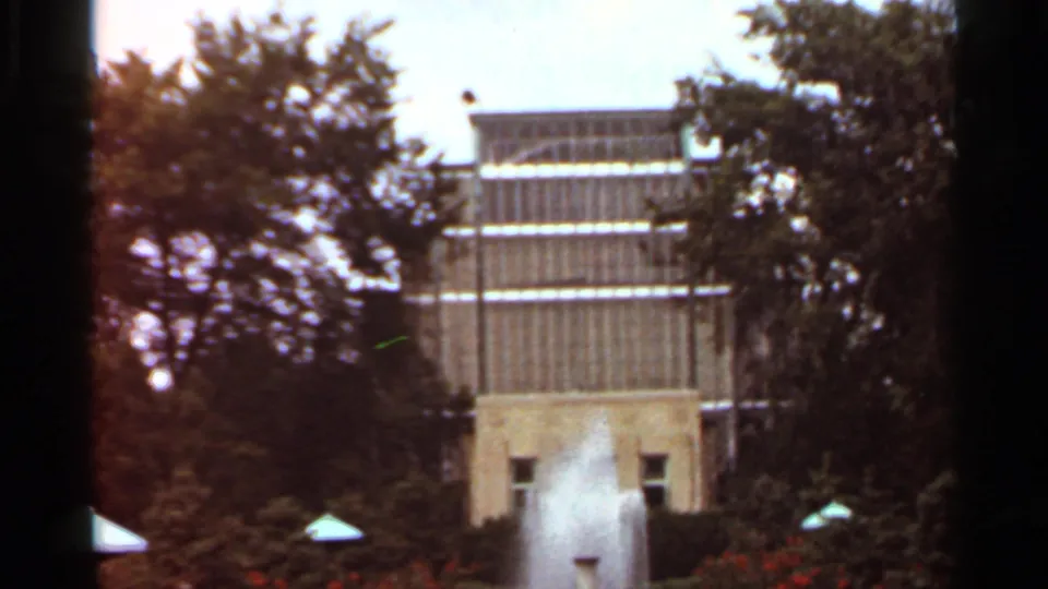 1966 Saint Louis frame with the fountain plume centered against a glass-fronted modernist building, trees, shrubs, and flower beds.