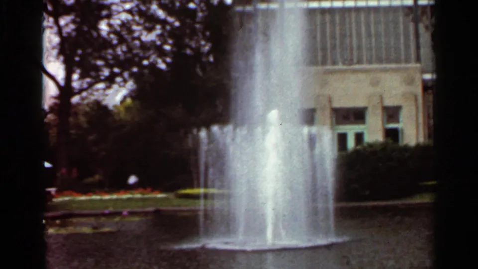 1966 Saint Louis frame showing a wider fountain pool view, with water jets, lawn, shrubs, trees, and a modernist building behind.