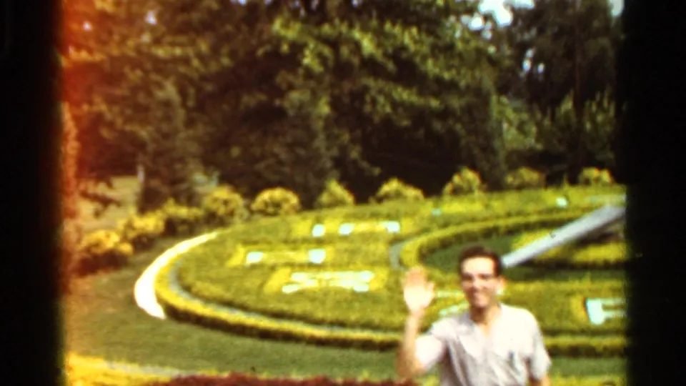 Adult man waves toward the 8mm camera beside a landscaped topiary clock in Saint Louis, Missouri, in 1966.