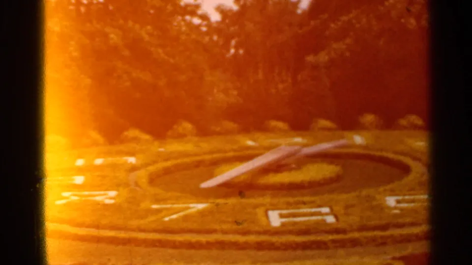 Close 8mm view of the floral clock design, with hedge borders, white numerals, and warm orange exposure across the frame.