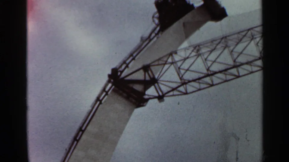 1966 Saint Louis 8mm frame looking up at exposed steel lattice and part of an unfinished concrete structure under cloudy sky.