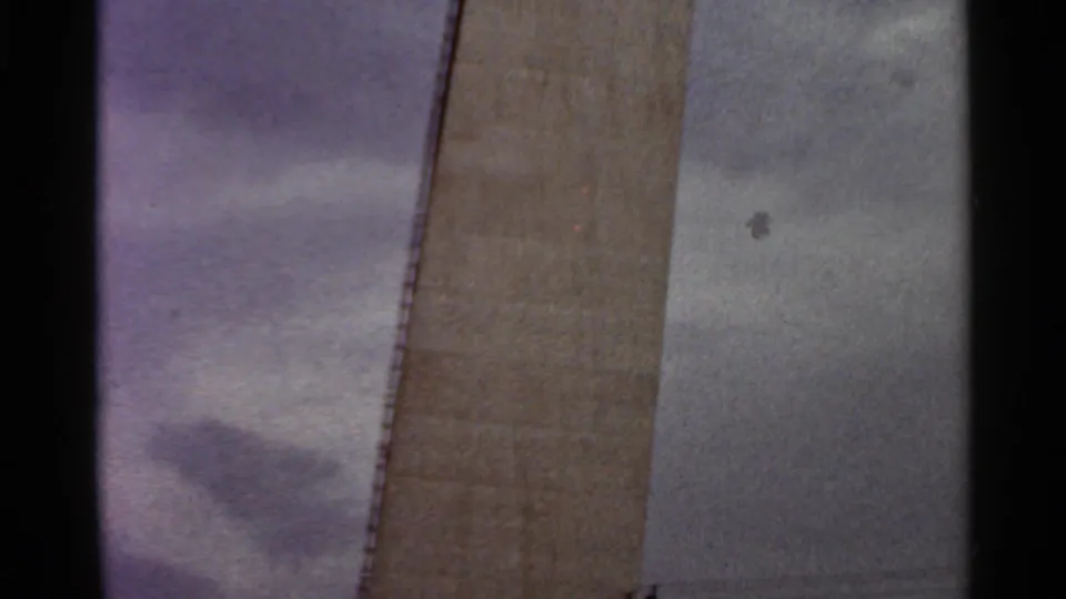 1966 Saint Louis home-movie frame with a tall concrete construction face and overcast sky.